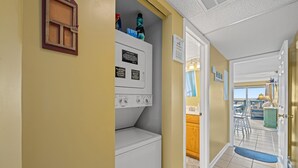 A laundry area with stacked washer and dryer, a window with a view, and a door leading to a balcony. - Nautical Watch 207-1BR/1BA Oceanfront condo in Windy Hill (North Myrtle Beach)