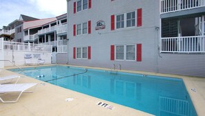 A swimming pool area with a pool, lounge chairs, and a building with red shutters and balconies.