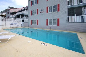 Uma área de piscina com piscina, espreguiçadeiras e um edifício com persianas vermelhas e varandas.
