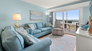 A living room with a blue sofa, a striped armchair, and a lamp. There is a painting on the wall and a view of the ocean through a sliding glass door. - Ocean Garden Villas C2-2BR/2BA Oceanfront Condo in Crescent Beach (North Myrtle Beach)