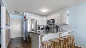 A modern kitchen with white cabinets, stainless steel appliances, a granite countertop, and wooden stools. - Ocean Garden Villas C2-2BR/2BA Oceanfront Condo in Crescent Beach (North Myrtle Beach)