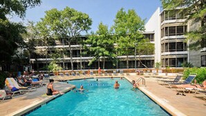 A swimming pool with a 7 FT depth marker, surrounded by lounge chairs and a building with multiple floors and large windows.