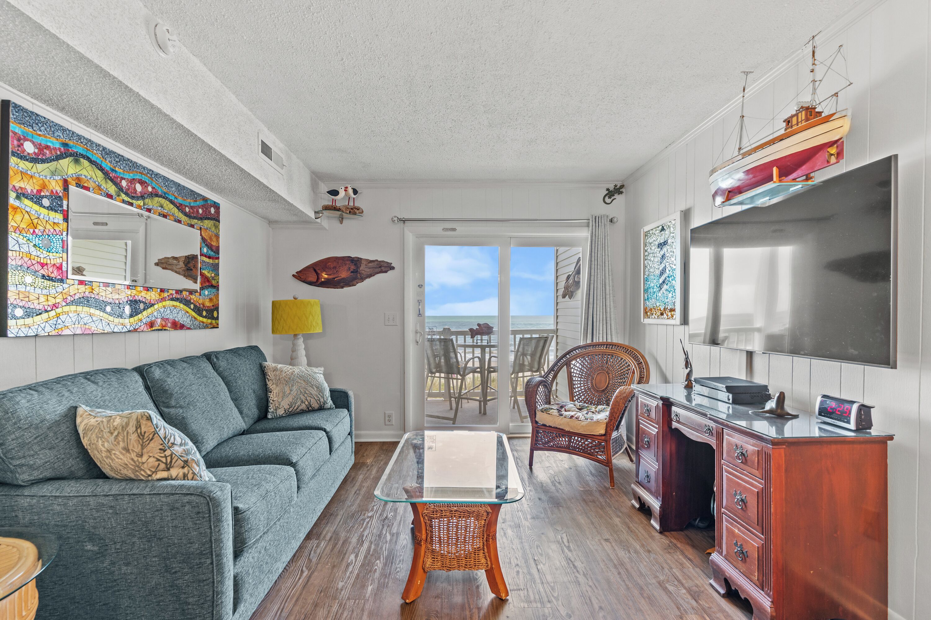 A living room with a sofa, a glass-top table, a wicker chair, a wooden cabinet, and a large window with a view of the ocean.