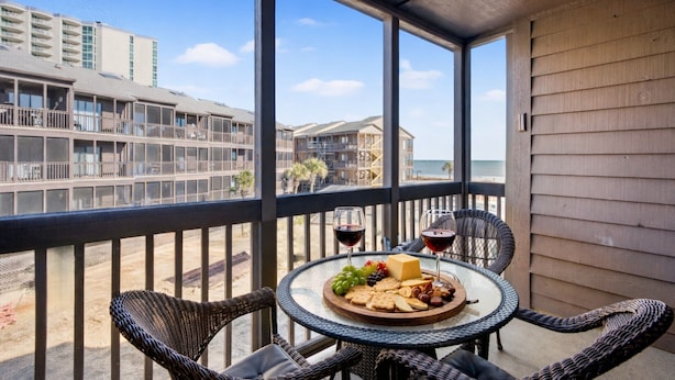 A balcony with wicker chairs, a round table with a cheese platter, wine glasses, and a view of buildings and the beach.