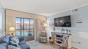 A living room with a sofa, armchair, and ottoman, a TV, a lamp, and a view of a building through a large window. - Tilghman Beach & Racquet Club 264-3BR/2.5BA Condo IN Ocean Drive (North Myrtle Beach)