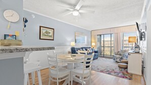 A modern living room with a dining area, a ceiling fan, and a large window. - Tilghman Beach & Racquet Club 264-3BR/2.5BA Condo IN Ocean Drive (North Myrtle Beach)