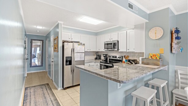 A modern kitchen with white cabinets, a granite countertop, and stainless steel appliances. There are two white barstools and a clock on the wall. - Tilghman Beach & Racquet Club 264-3BR/2.5BA Condo IN Ocean Drive (North Myrtle Beach)