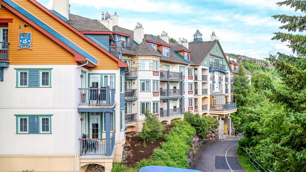 A row of colorful multi-story buildings with balconies, a narrow road, and lush greenery. - The MountainSide by Escapades Tremblant (Mont-Tremblant)