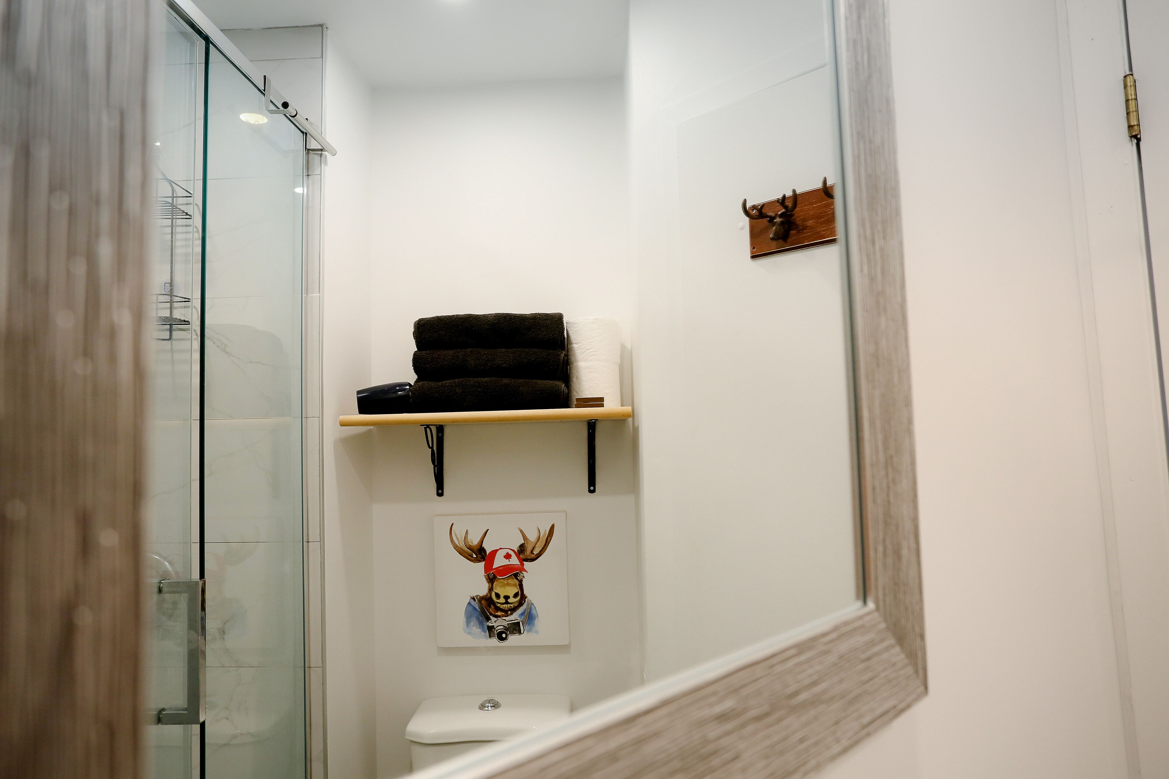 A bathroom with a glass shower enclosure, a toilet, a towel rack, a shelf with a towel, and a wall-mounted picture of a deer wearing a red cap.