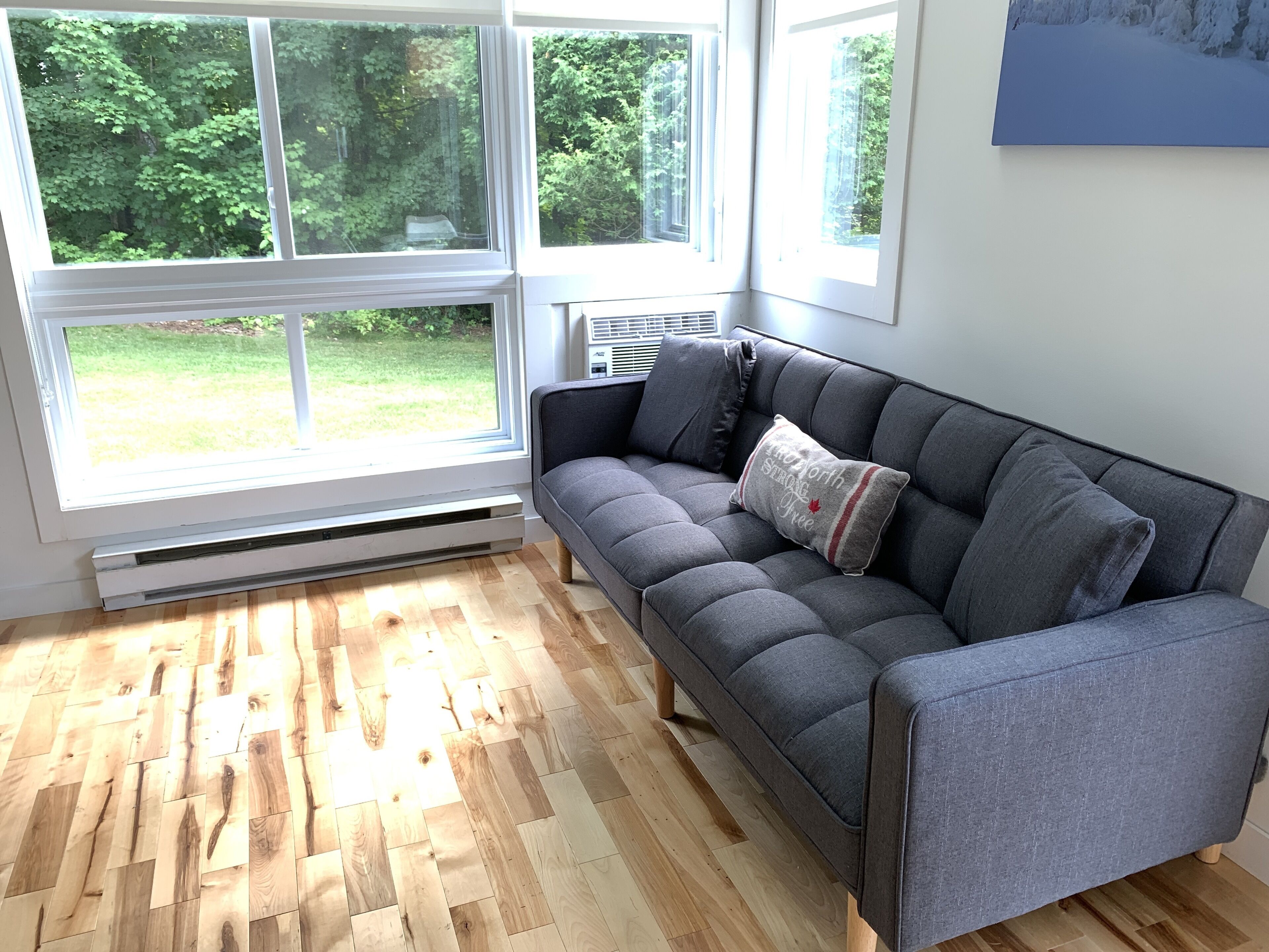 A modern living room with a grey sofa, a large window with a view of greenery, and a wooden floor.