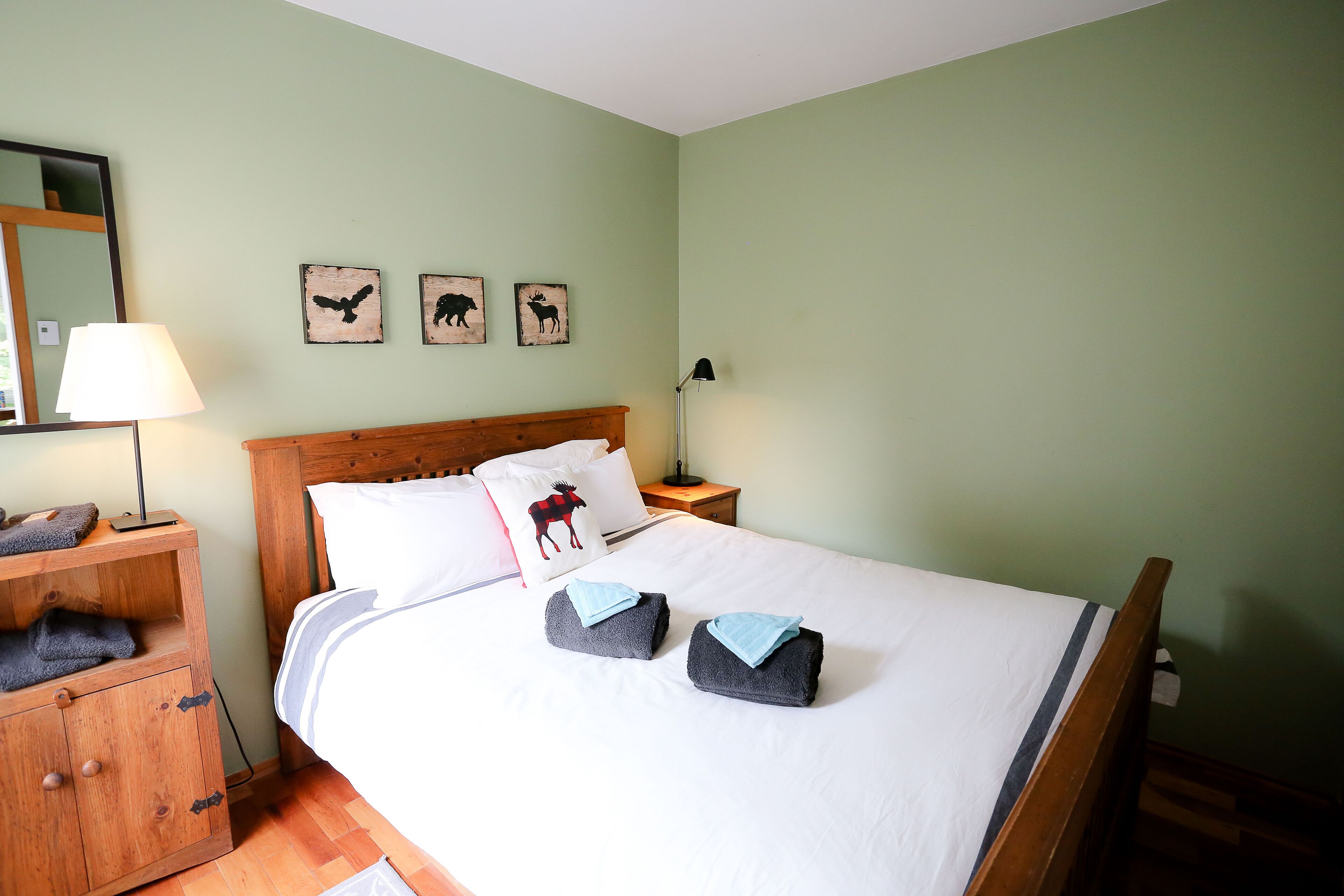 Un lit bien fait avec deux serviettes, une table de chevet en bois avec une lampe, un miroir et trois œuvres d’art murales sur le thème des animaux.