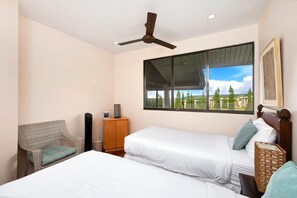 Un dormitorio con dos camas, un ventilador de techo, una ventana con vista a edificios y una silla de mimbre.