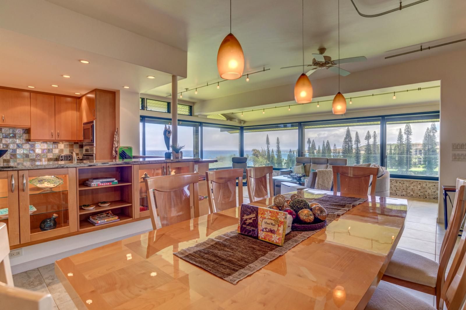 A modern kitchen with wooden cabinets, a dining area with a glass table, and large windows offering a view of the outdoors.