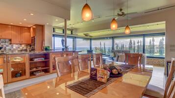 A modern kitchen with wooden cabinets, a dining area with a glass table, and large windows offering a view of the outdoors.