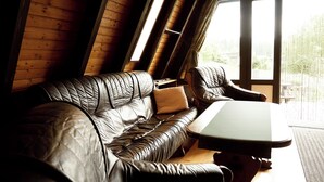 A wooden interior with a leather sofa and armchair, a small table, and large windows offering a view of greenery outside.