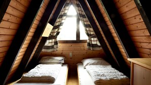 A small, triangular attic bedroom with two beds, wooden walls, and a window with curtains.