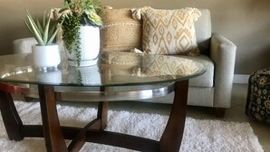 A round glass coffee table with dark wooden legs, a white textured rug, a grey sofa with patterned cushions, and two potted plants. - Luxury Condo - Tranquil Desert Retreat  (Palm Desert)