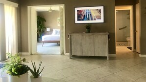 A spacious living area with a large bed, a TV mounted on the wall, and a potted plant on a wooden console table. - Luxury Condo - Tranquil Desert Retreat  (Palm Desert)
