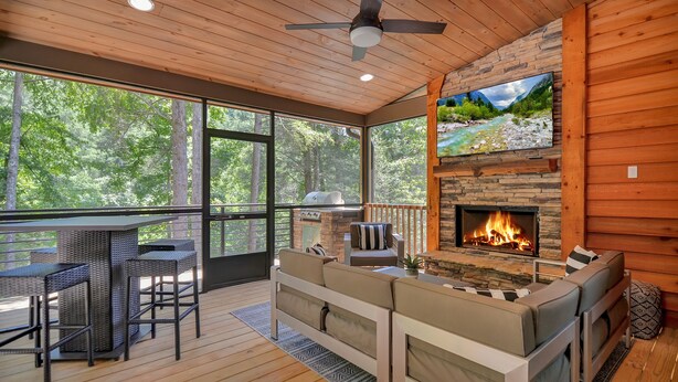 A wooden deck with a stone fireplace, outdoor seating, and a large window overlooking a forest. - Watersong Falls ~ Private Waterfall, Hot Tub, Fire Pit & More (Ellijay)