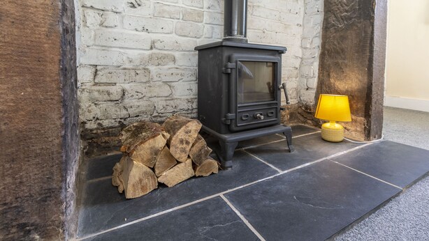 A wood-burning stove next to a white brick wall, with a stack of firewood and a yellow lamp on the floor.