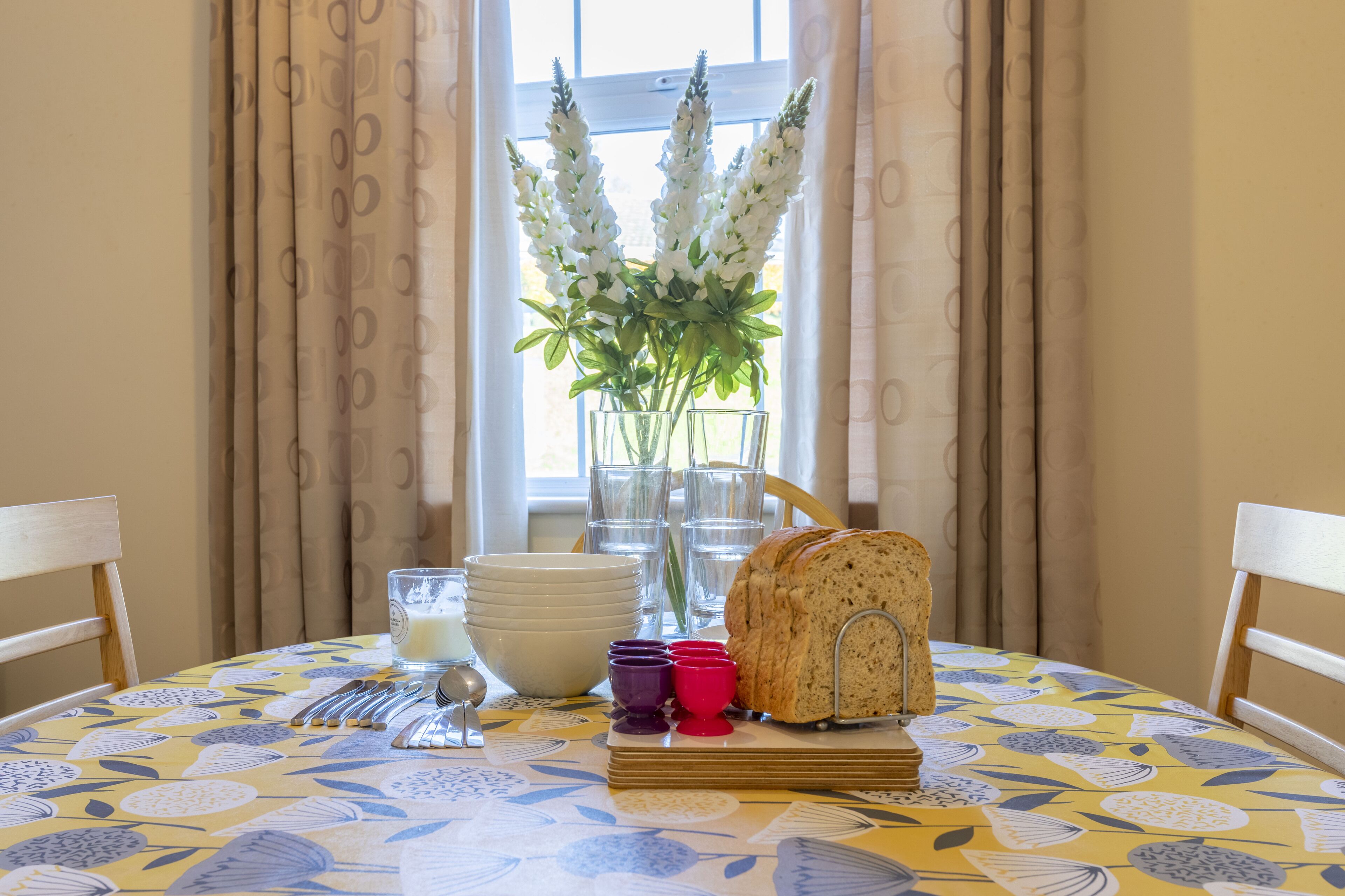 Une table à manger recouverte d’une nappe à motifs floraux, un pain, un vase rempli de fleurs et une fenêtre avec des rideaux.