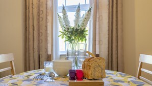 A dining table with a floral tablecloth, a loaf of bread, a vase with flowers, and a window with curtains.