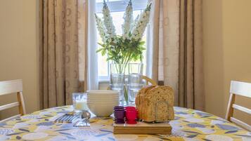 Une table à manger recouverte d’une nappe à motifs floraux, un pain, un vase rempli de fleurs et une fenêtre avec des rideaux.