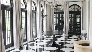 Una habitación con piso a cuadros blanco y negro, ventanas amplias con cortinas, un candelabro y una mesa redonda.