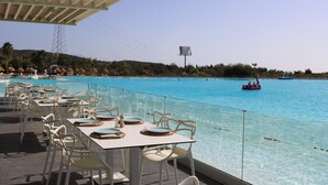 Ein Essbereich am Pool mit Tischen und Stühlen, mit Blick auf ein klares, blaues Schwimmbecken und ein Boot in der Ferne.