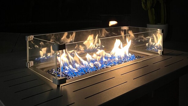 Ein rechteckiges, freistehendes Feuerbecken mit Glasabdeckung, umgeben von einer dunklen Terrassenfläche mit einem Kaktus und Lichterketten im Hintergrund.