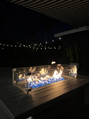 A rectangular outdoor fire feature with a glass enclosure, surrounded by a dark patio area with a cactus plant and string lights in the background.
