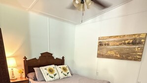 Une chambre à coucher avec un lit en bois, une table de chevet avec une lampe, un ventilateur de plafond et un tableau représentant un paysage accroché au mur.