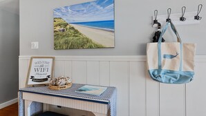 Un tableau représentant une plage, accroché au mur, une pancarte de bienvenue, une table avec un panier et un livre, et un sac fourre-tout suspendu.