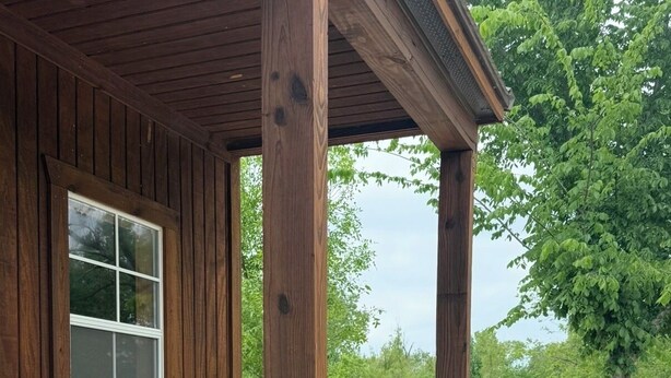 Une véranda en bois avec un fauteuil à bascule et un banc, une fenêtre, et une vue sur un espace verdoyant avec des arbres.