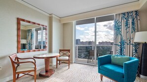 A hotel room with a large window, a round table with two chairs, a blue armchair, and a mirror on the wall.
