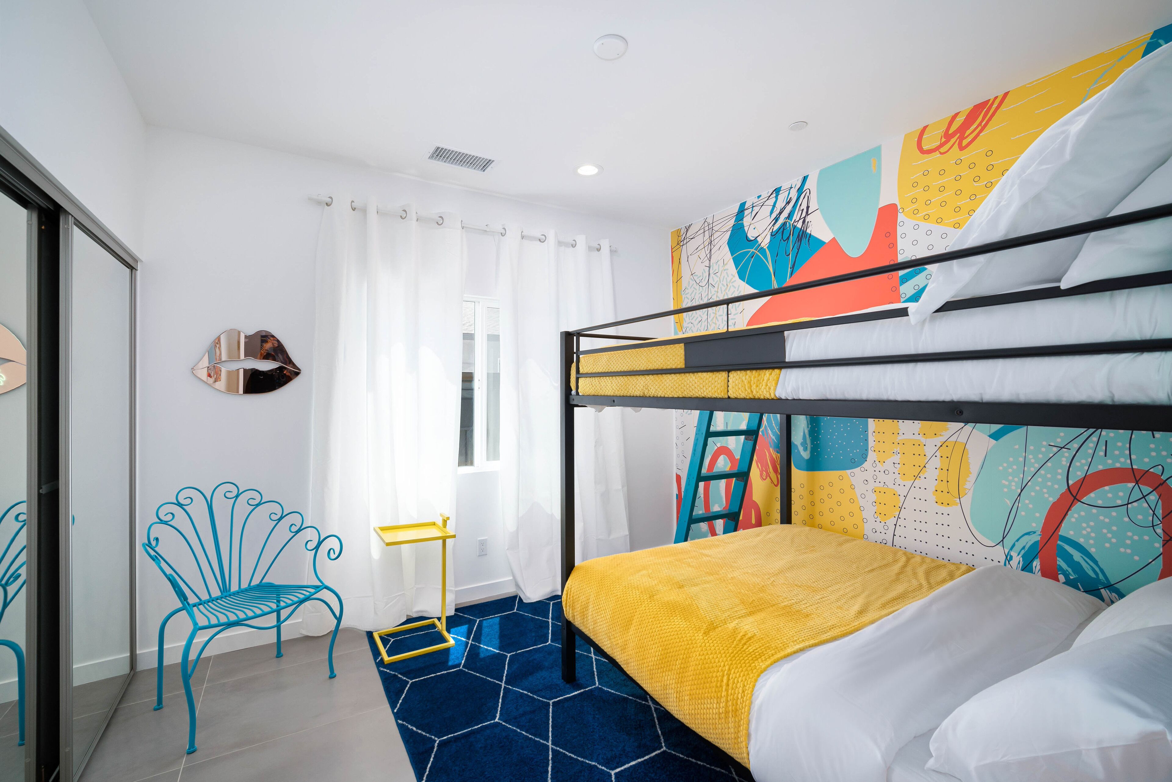 A room with a bunk bed, a blue chair, a small table, and a colorful abstract wall art.