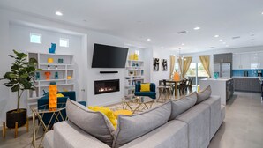 Ein modernes Wohnzimmer mit einem grauen Sofa, einem Kamin und einem Fernseher. Es gibt eine Küchenzeile mit Schränken und einem Kühlschrank.