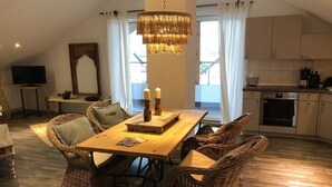 Un coin repas avec une table en bois et des chaises en osier, un lustre et un téléviseur en arrière-plan.