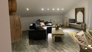 Un salon moderne de style industriel, avec un plafond haut, un canapé noir, une table basse en bois et un fauteuil en osier.