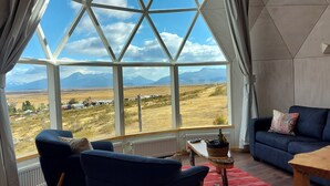 A room with a large window offering a view of a mountainous landscape. There is a blue sofa, a wooden table, and a radiator. - Geodesic Dome with Stunning Glacier and Mountain views (Puerto Natales)