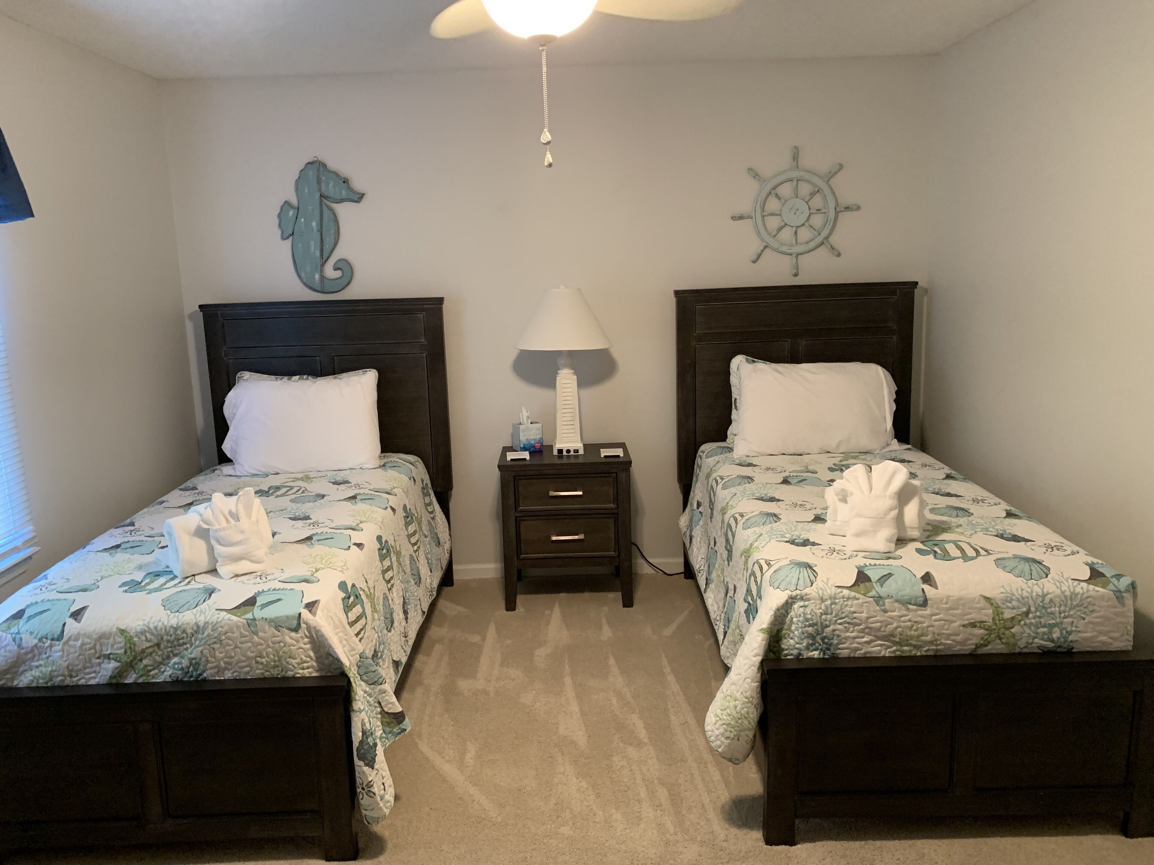 Habitación de hotel con dos camas, cada una decorada con un adorno de caballito de mar, una rueda de barco y una mesita de noche con lámpara y pañuelos.