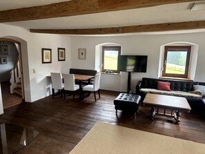 A spacious living area with a wooden floor, a dining table with chairs, a sofa, a television, and a staircase.