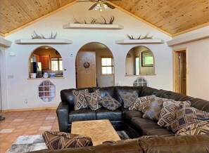 A spacious living room with a large black leather sofa, patterned throw pillows, and a wooden coffee table. The room features a high wooden ceiling with a fan, and there are three arched windows with decorative antler motifs above them. - Private Haven+5 Acres+Sprawling Yards+Spectacular Waterfall Koi Pond!  3BR/2Bath (Bayfield)