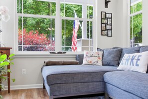 Une pièce avec un canapé gris, une fenêtre donnant sur des arbres et un drapeau américain, et un mur orné de tableaux encadrés.