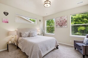 Un dormitorio con cama, mesita de noche, lámpara y dos ventanas con vistas a zonas verdes.