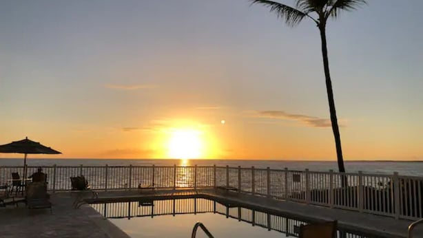 A sunset view from a poolside area with lounge chairs and a palm tree.