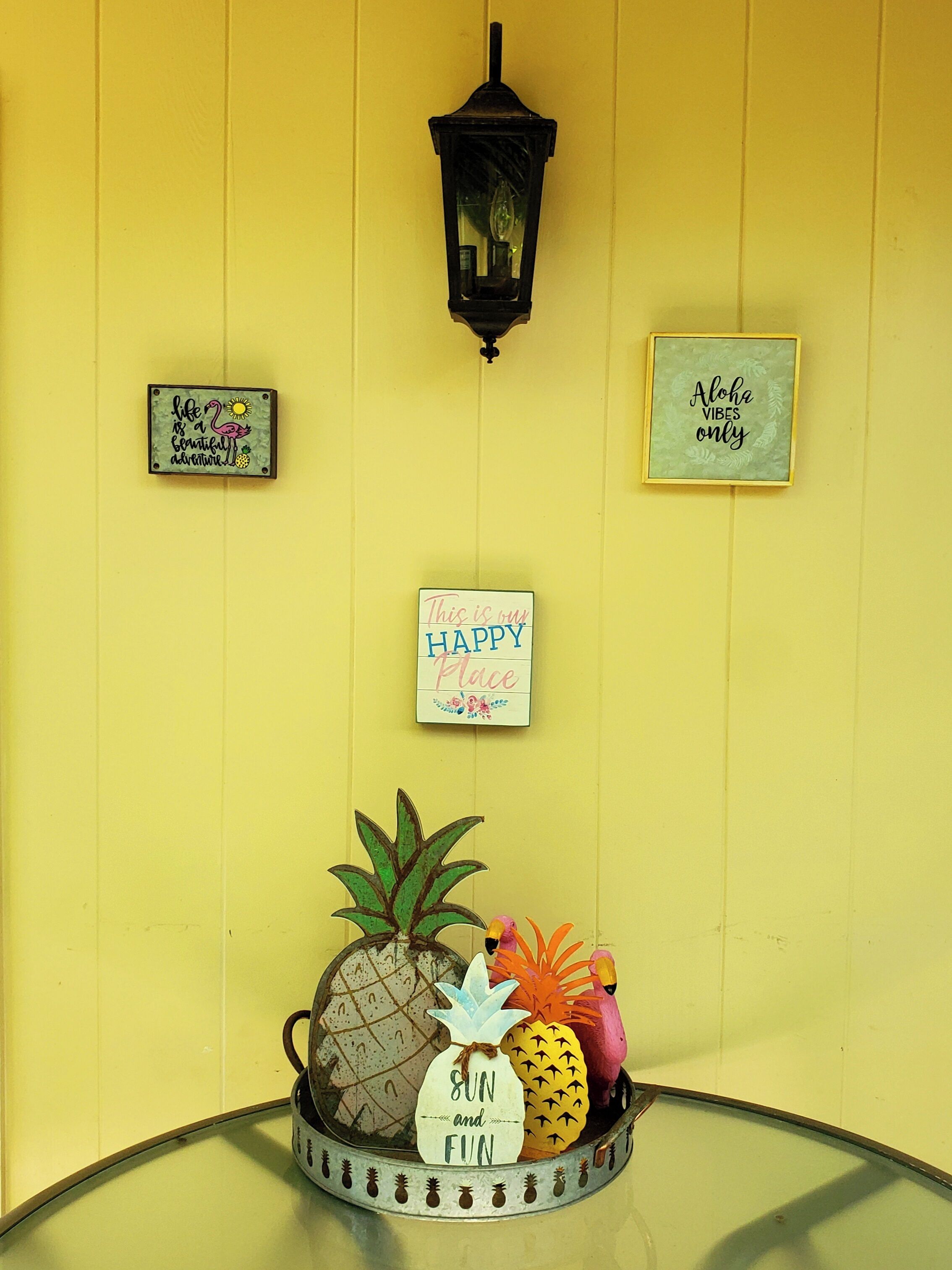 Una pared amarilla con tres letreros: “life is a beautiful adventure”, “This is our happy place” y “Aloha vibes only”. Abajo, una bandeja contiene una piña, un flamenco y una planta.