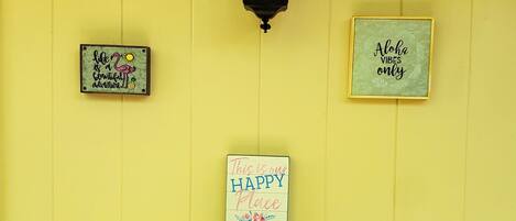 Una pared amarilla con tres letreros: “life is a beautiful adventure”, “This is our happy place” y “Aloha vibes only”. Abajo, una bandeja contiene una piña, un flamenco y una planta.