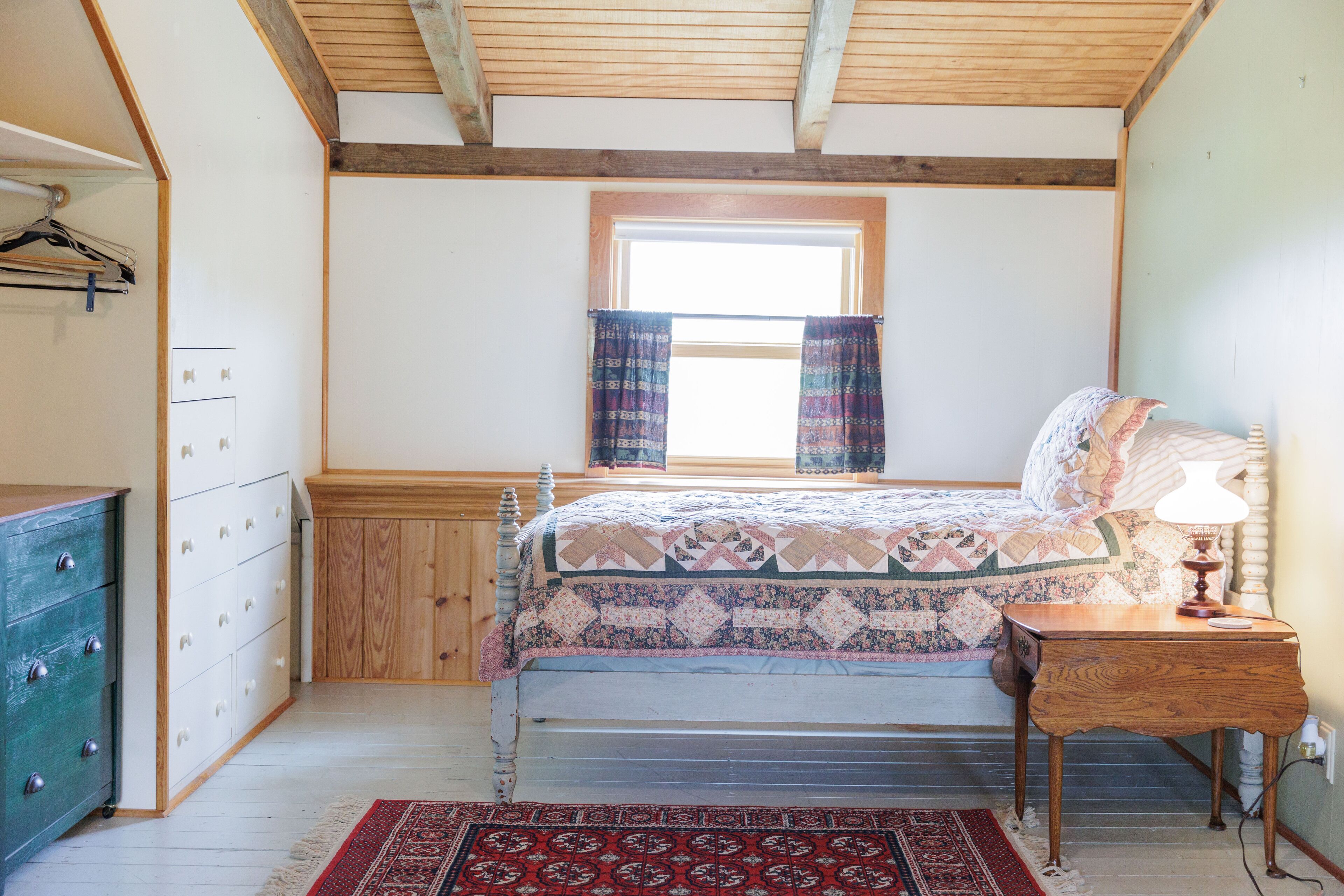 A bedroom with a bed, wooden headboard, a nightstand with a lamp, a dresser, a window with curtains, and a patterned rug.
