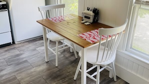 Une cuisine avec une table en bois, des chaises blanches, un réfrigérateur blanc et une fenêtre avec des stores.